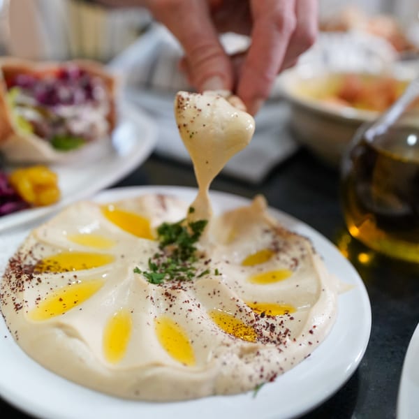 A hand dips pita bread into a plate of hummus drizzled with olive oil and spices.