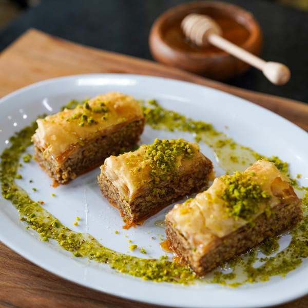 Three pieces of baklava with honey and crushed pistachios on a white plate, with a honey dipper in the background.