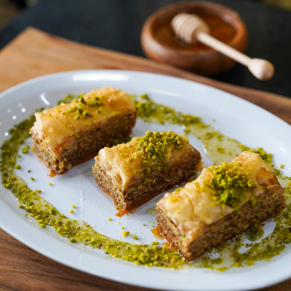Three pieces of baklava with honey and crushed pistachios on a white plate, with a honey dipper in the background.